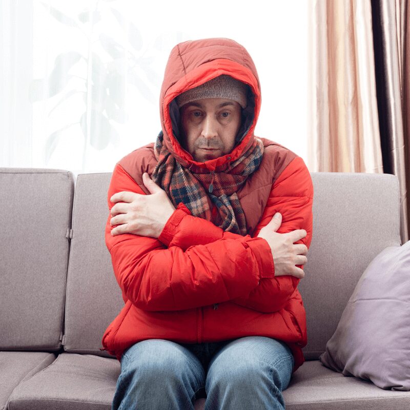 middle-age-male-model-in-red-parka-and-checkered middle-aged white man sitting in winter coats on his couch because he’s cold.